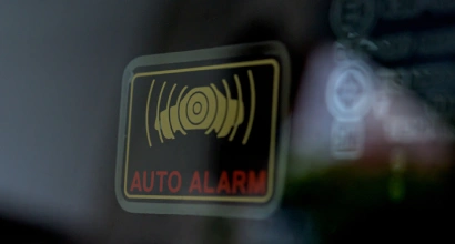 Close-up of a car window sticker showing a yellow symbol with sound waves and the words "AUTO ALARM" in red text.