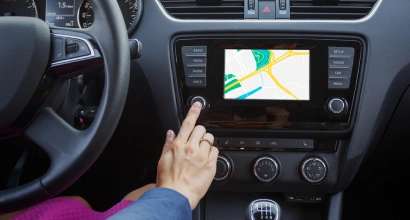 Driver pressing a button on a car's touchscreen navigation system displaying a colorful map, with dashboard controls and steering wheel visible.