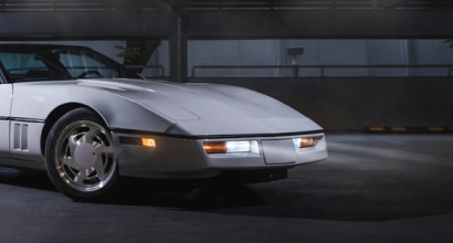 Close-up of the front end of a silver vintage sports car with headlights on, parked in a dimly lit garage.