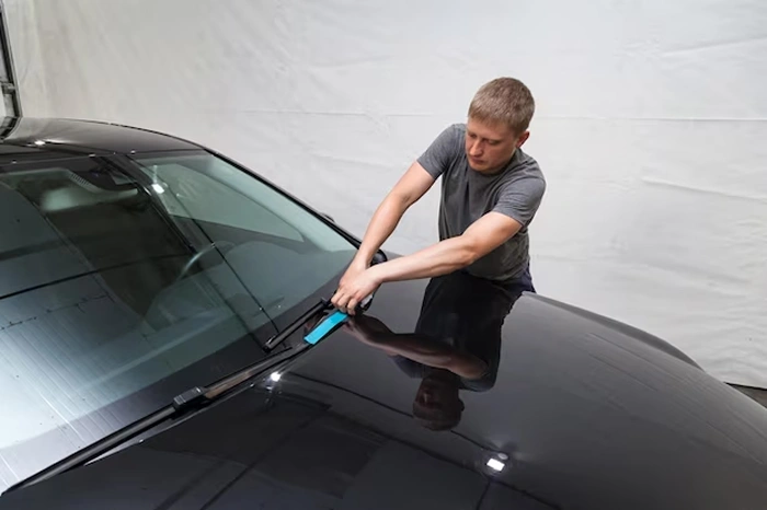 Man in a gray t-shirt uses a blue squeegee to apply or smooth a film on the windshield of a black car in a workshop setting.