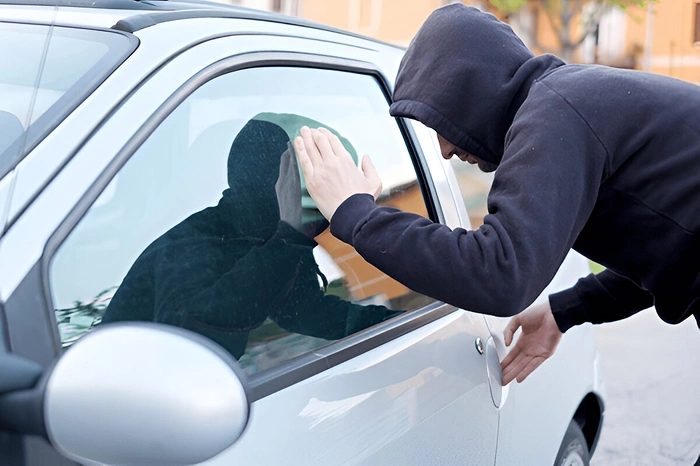 Person in a black hoodie peering into the window of a parked car while gripping the door handle, suggesting suspicious or attempted unauthorized entry.