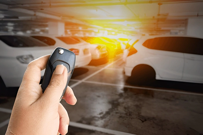 Hand holding a car remote key fob, aimed at a row of parked white cars in a dimly lit garage with bright sunlight shining in the background.