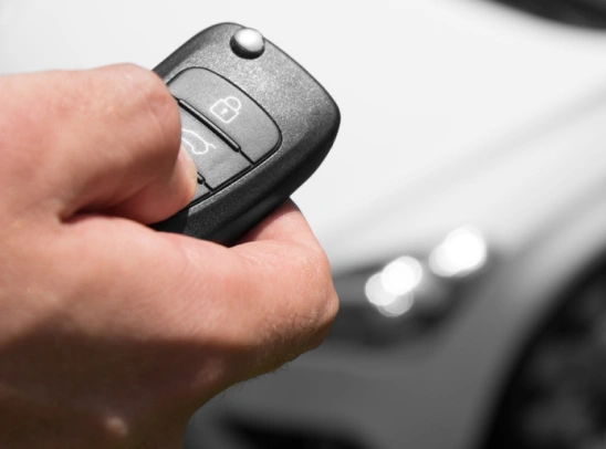 Hand pressing a button on a black car key fob with lock and trunk icons, with a white car blurred in the background.