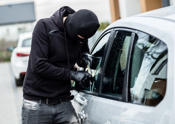 Masked individual in dark clothing attempting to break into a silver car using a tool, indicating a car theft in progress.