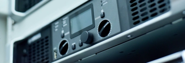 Close-up of professional audio equipment mounted in a rack, featuring control knobs, a display screen, and ventilation panels.