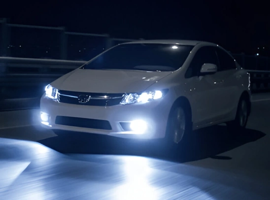 White Honda sedan driving at night with bright white LED headlights and fog lights illuminating the road ahead.