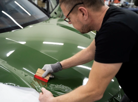 Man applies a green vinyl wrap to a car hood using a squeegee tool, smoothing the material with precision while wearing a protective glove.