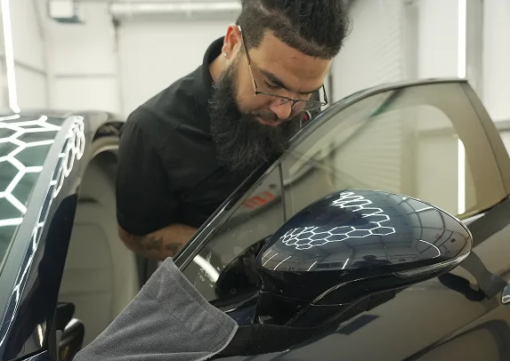 Man with glasses and a beard closely inspects the surface of a black car door near the side mirror, with a hexagonal light reflection visible on the paint.