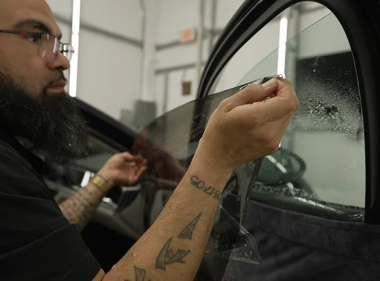 Bearded man with tattoos carefully applies a tinted film to a car window inside a workshop, with the glass slightly wet for installation.