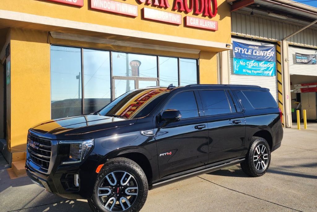 A shiny black GMC SUV with dark-tinted windows is parked outside Extreme Car Audio, a shop offering window tinting, lighting, and car alarms. The storefront has bold signage and banners promoting no-credit-needed financing and customization services.