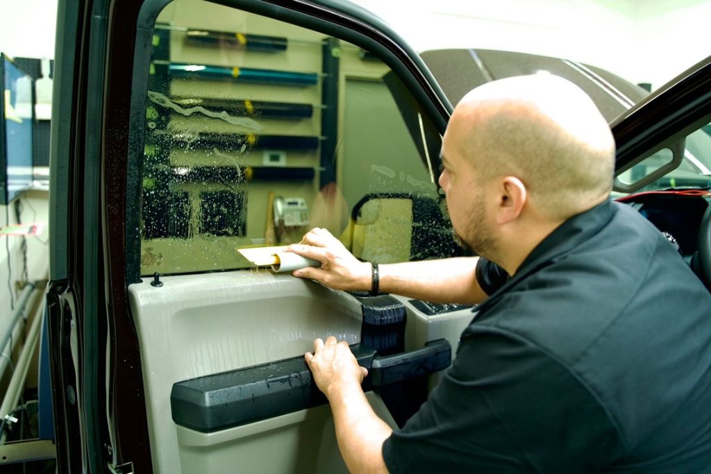 A technician uses a squeegee to smooth window tint film onto a vehicle’s side window in a clean workshop. Water and application solution are visible on the glass, indicating the precision involved in professional tint installation.