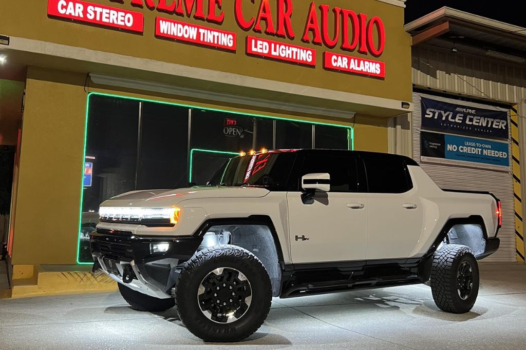 A white Hummer EV with dark tinted windows is parked at night in front of Extreme Car Audio, a storefront with bright neon and red signage offering window tinting, car stereo, LED lighting, and car alarms.