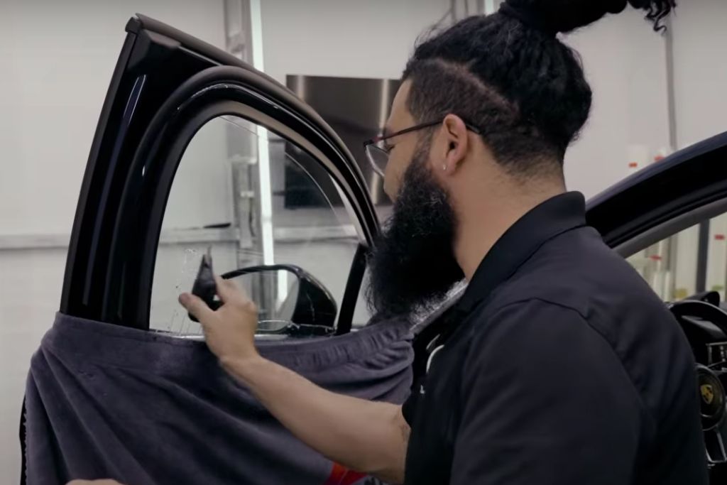 A technician with a beard and tied-back hair sprays solution on a car window in preparation for applying tint film, with a protective towel draped over the door frame inside a clean, well-lit shop.