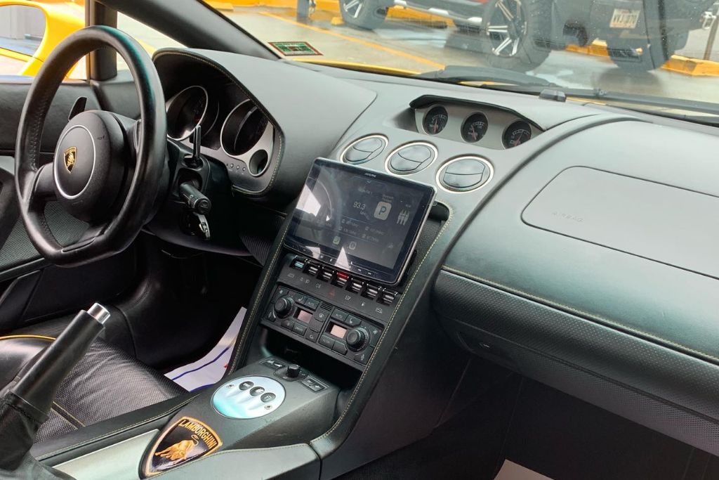 Interior view of a Lamborghini showing the dashboard, center console, and steering wheel, with visible touchscreen controls and tinted windows allowing in diffused light from the outside parking lot.