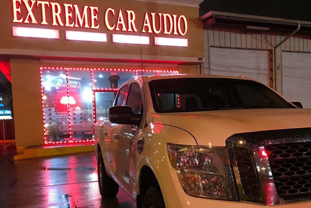 A white pickup truck with tinted windows is parked in front of Extreme Car Audio at night, with the shop’s bright red neon signage and window lighting reflected on the wet pavement.