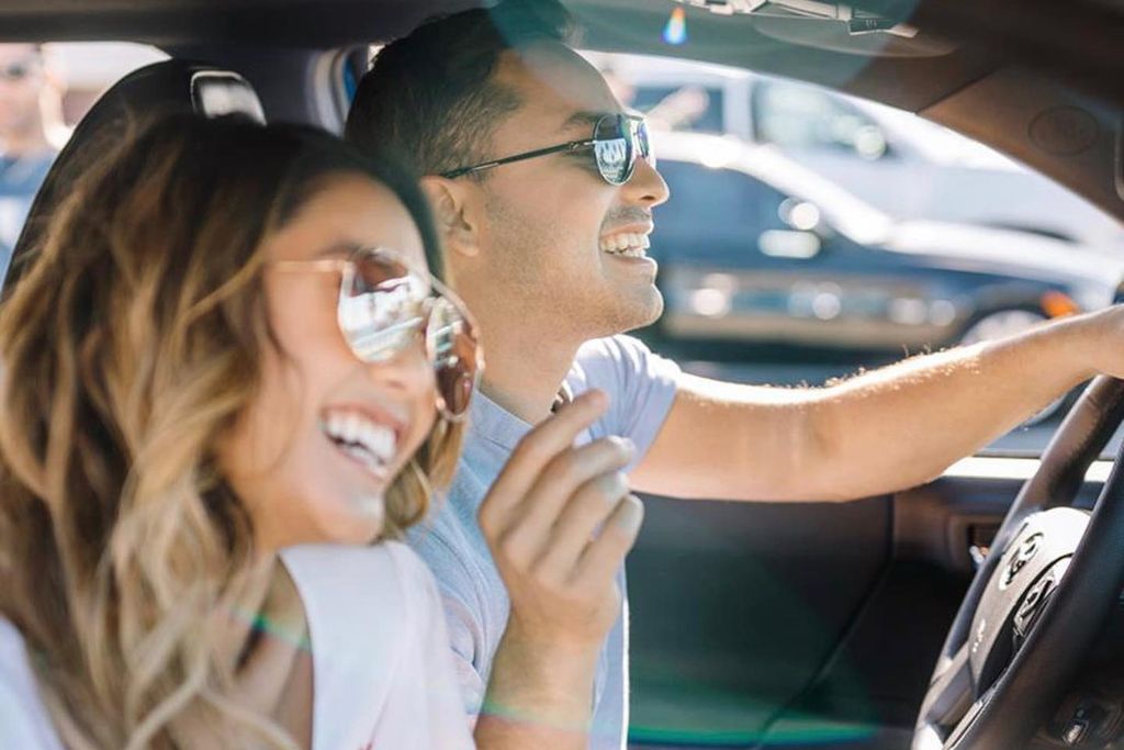 A smiling couple wearing sunglasses enjoys a sunny drive inside a car. Bright sunlight pours through the windshield, suggesting the importance of heat-rejecting tint for comfort and protection in sunny climates.