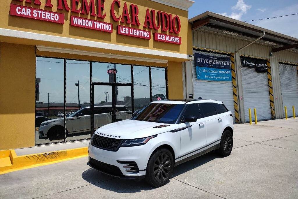 A white Range Rover SUV with dark tinted windows parked in front of Extreme Car Audio, a shop offering services like window tinting, LED lighting, and car alarms, under a bright sunny sky.