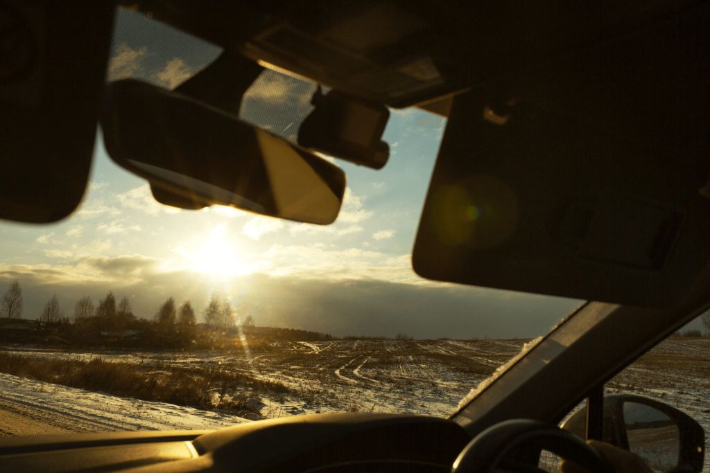 View from inside a car looking out through the windshield at a low winter sun over a snowy field, with the sun’s glare softened by the tinted glass and the driver’s silhouette partially visible.