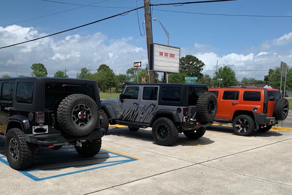 Three off-road vehicles with dark tinted windows—two Jeep Wranglers and one orange Hummer—are parked in bright sunlight in a lot near a street intersection, beneath a mostly clear sky.