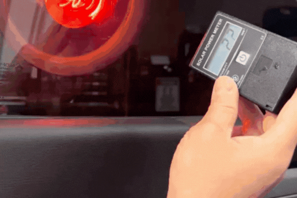 A hand holds a digital solar power meter reading "22" in front of a tinted car window, demonstrating how the tint significantly blocks solar energy. The background shows a glowing red heat lamp shining on the glass, emphasizing the window tint's heat-reducing effect.