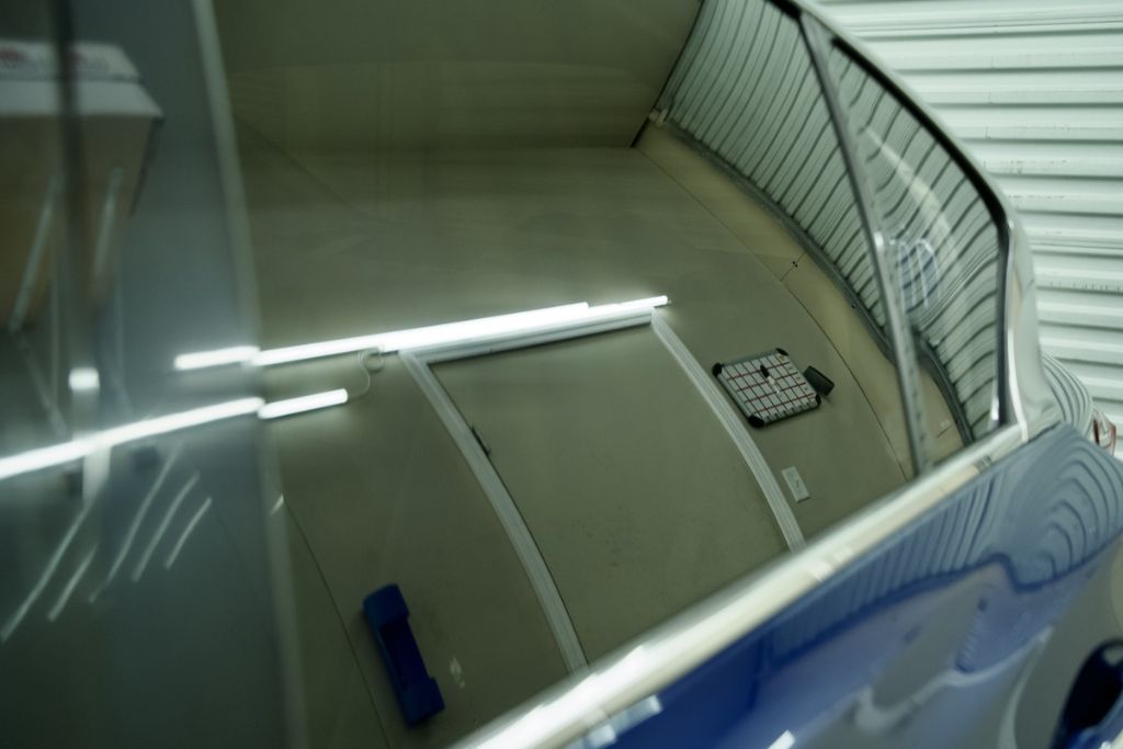 Close-up of a tinted car window reflecting fluorescent ceiling lights and a garage wall, showcasing the smooth, clear finish of a high-performance ceramic window tint.