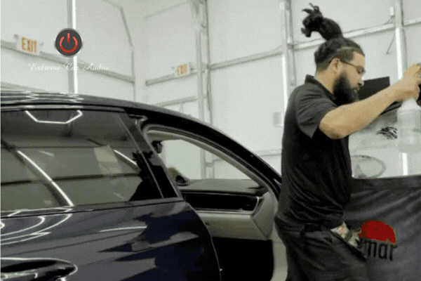 A technician at Extreme Car Audio preps a luxury vehicle’s window for tint installation, spraying the surface with solution inside a clean, well-lit shop. The motion highlights precision and care in the professional process.