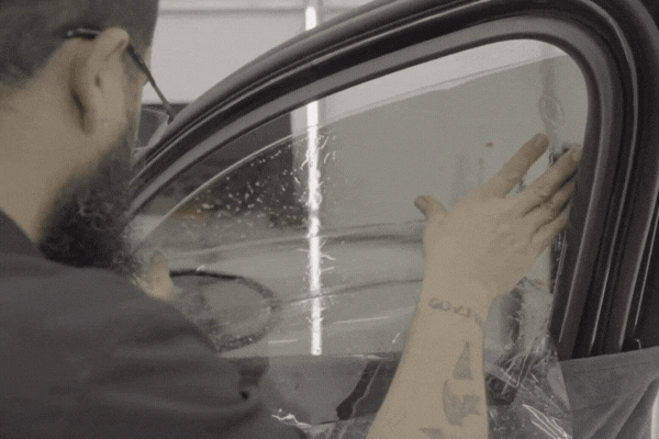 A bearded technician carefully smooths window tint film onto a car’s side window, ensuring a clean and even application. The image supports the topic What Affects the Lifespan of Window Tint? by illustrating the importance of professional installation for long-lasting results.