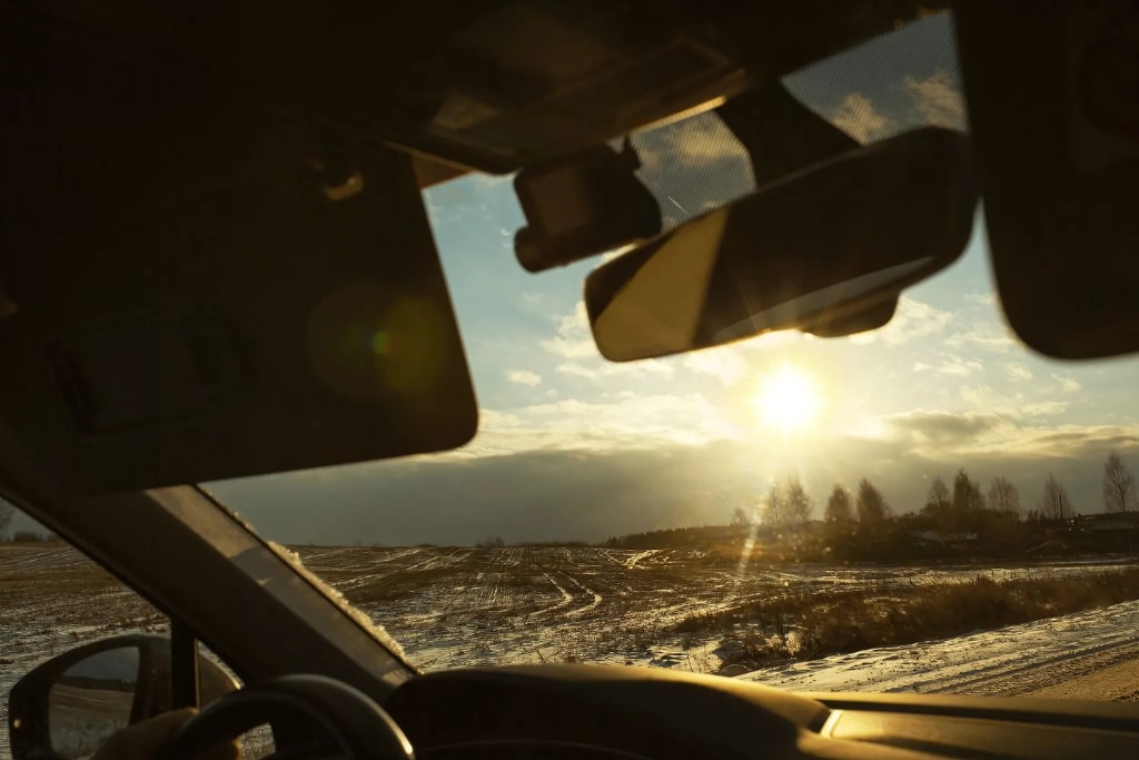 View from inside a car looking out through the windshield at a low winter sun over a snowy landscape, illustrating the intense sunlight exposure that contributes to window tint deterioration. The image supports the topic Does Tint Deteriorate Over Time by visually emphasizing UV and heat exposure.